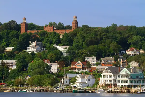 View of a small town with houses along the waterfront, and a castle-like structure surrounded by greenery in the background