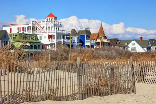 Sand dunes with a wooden fence in the foreground, a row of colorful Victorian-style houses in the background