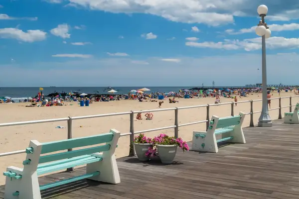 A boardwalk with benches, lampposts, and a beach with people and umbrellas in the background