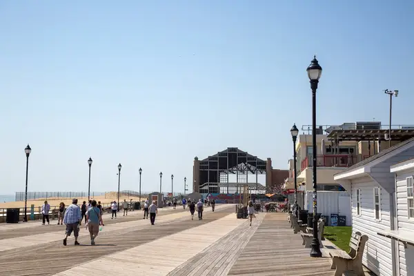 A boardwalk scene with people walking, historic structure in the distance, streetlamps, and shops along one side in Asbury Park, New Jersey