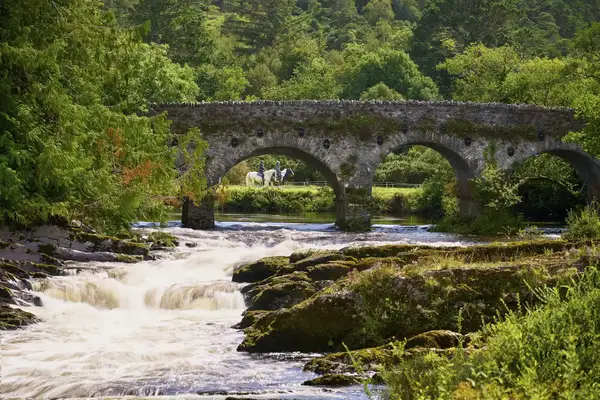 A stone bridge over a stream with a person horseback riding visible in the distance