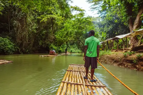 A man standing on a bamboo raft gliding through a lush tropical river landscape