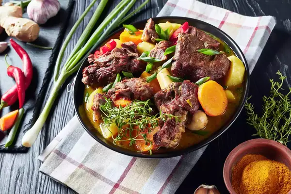 A plate of stew with vegetables, displayed on a checkered napkin and surrounded by spices and ingredients on a dark background