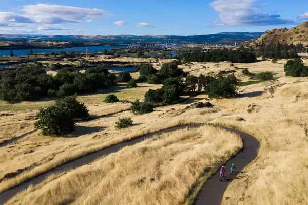 Cyclists on a winding path through a dry, open landscape with hills and sparse vegetation, the Columbia River visible in the background