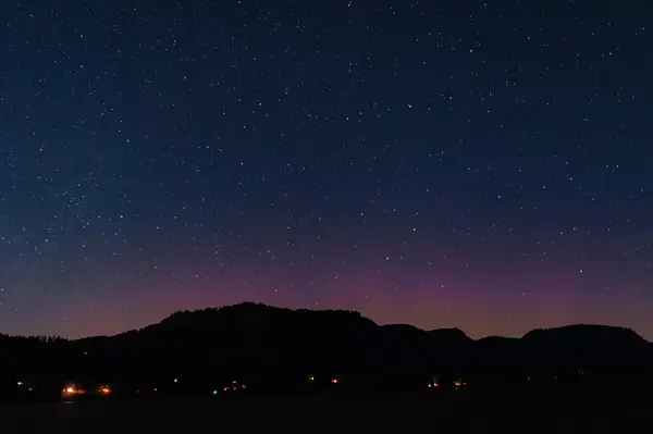 Starry night sky over a silhouette of mountains with scattered lights below