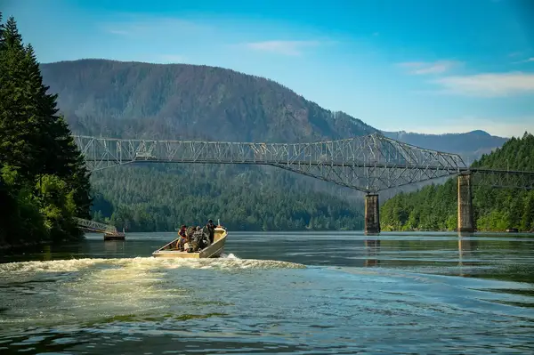 People on a motorboat traveling under a suspension bridge spanning a river, with hills in the background