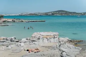 A person sunbathing on a rocky beach near the sea, with distant hills and a town visible along the shoreline