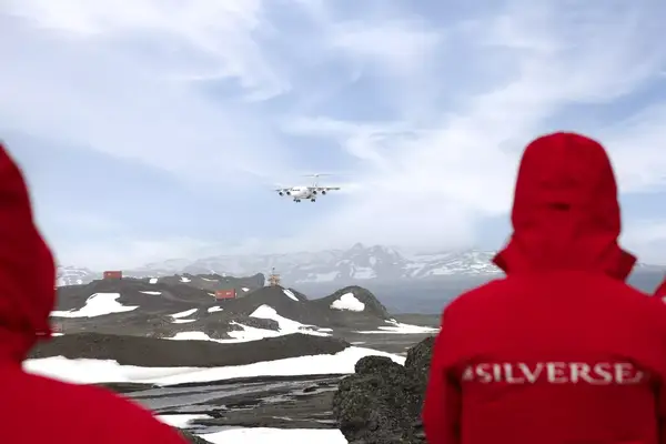 People in red jackets watch a plane approacing a gravel runway