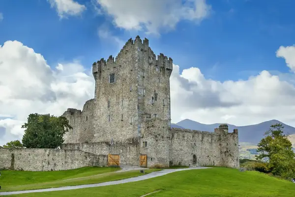 A stone castle with a surrounding wall situated on a grassy landscape with mountains in the background