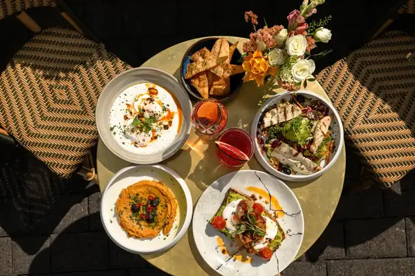 Outdoor cafe table with plated dishes such as bread salads and dips with drinks alongside decorative flowers