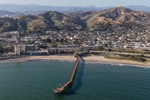 Aerial view of a coastal city with a long pier extending into the ocean hills visible in the background beach and buildings in foreground