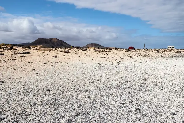 Landscape featuring a white pebblestrewn beach with a mountain in the background and a few parked vehicles in the distance