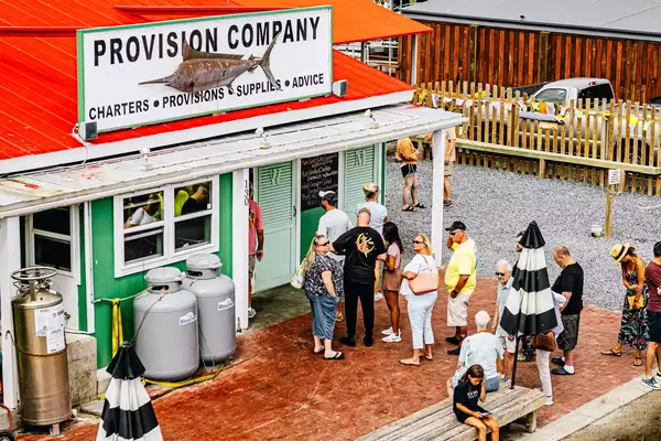 A line of people waiting for tables forms in front of Provision Company located on the Southport Waterfront. Intercoastal waterway in background with American and North Carolina flag flying.