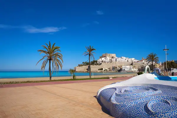A curvy sculptural bench covered in blue mosaic tiles sits in the foreground of this shot of the boardwalk and beach in Peniscola, Spain, with a castle-topped hillside town in the background.