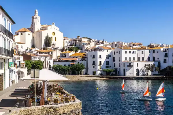 Whitewashed, tan-roofed buildings and a large white church surround a small blue harbor in Cadaques, Costa Brava.