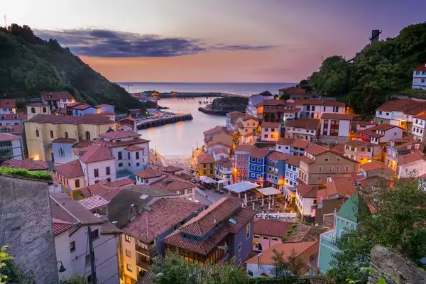 An aerial or hilltop photo looks down on the village of Cudillero at sunset, with a series of colorful, tile-roofed buildings surrounding a central square and a harbor that opens up to the sea, with an orange and purple sunset beyond.