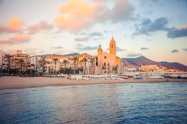The Church of Sant Bartomeu and Santa Tecla stands tall and pink-hued in the sunset looking in from the water towards Sitges, Spain.