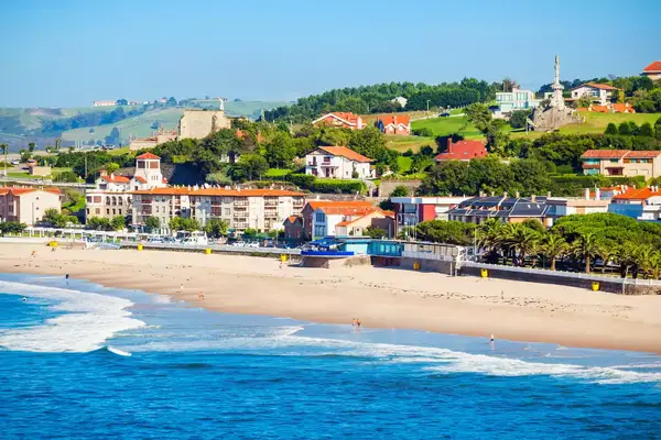 An aerial panoramic view shows the city beach of Comillas, Cantabria, Spain, which backs u to a stretch of red-roofed buildings and a rolling green hill.