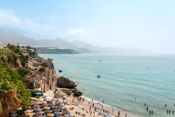 Crowded beach with people swimming and sunbathing, cliffs and scattered umbrellas nearby, calm sea stretching into the horizon.