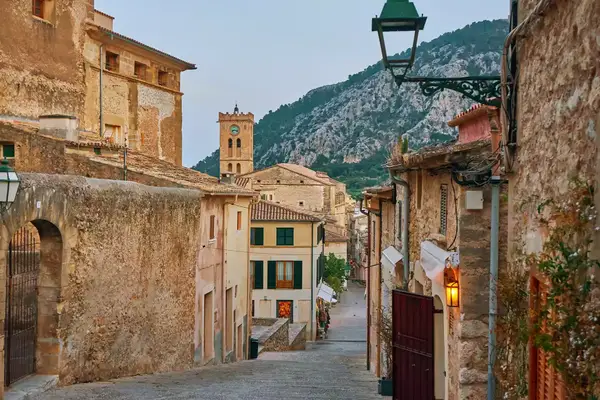A view down the Carrer del Calvari in Pollenca, Mallorca, featuring old weathered stone houses, a square church steeple in the distance, and rocky mountains beyond.