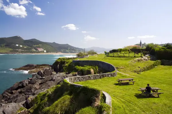 A green lawn studded with picnic tables and bordered by a curving stone wall sits just above the rocky seashore in Mundaka, Basque Country.