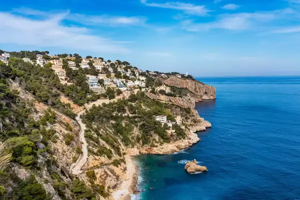 A hillside town covered with trees, rocks, and white buildings sits beside Javea Playa Ambolo beach in Xabia, Alicante, Spain.