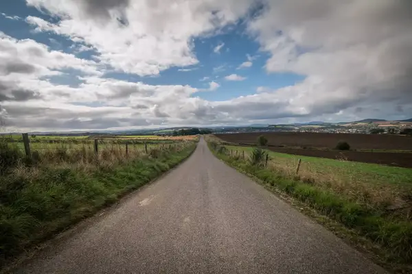A straight rural road stretching into the distance surrounded by fields and crops, under a cloudy sky