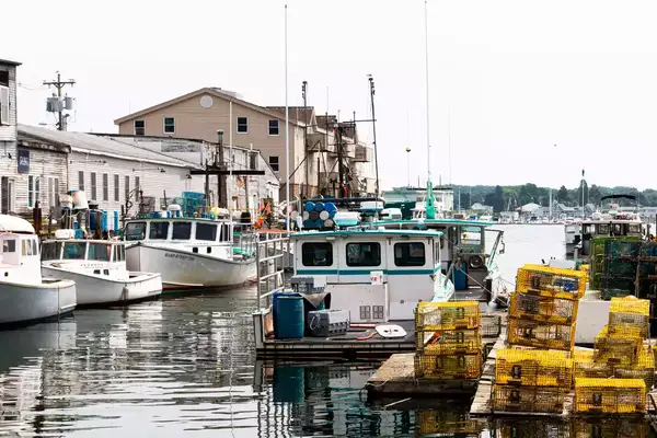 Commercial fishing boats docked in a canal with colorful lobster traps stacked on the dock behind buildings.
