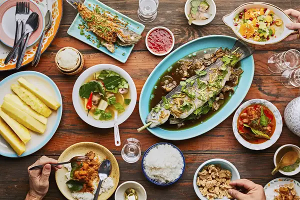 A table arranged with a variety of dishes including roasted fish rice and fruits with hands visible serving and eating the meal