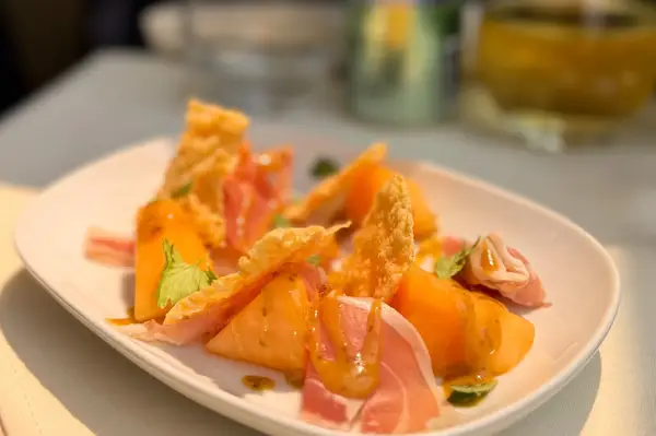 A plated dish featuring thinly sliced ham melon pieces and crispy topping on a white plate