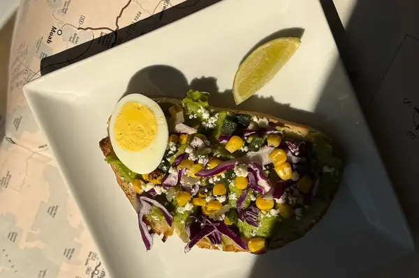 Avocado toast topped with sliced egg corn lettuce cabbage and cheese served with a lime wedge on a white plate