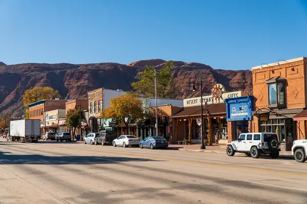 A downtown street scene in Moab featuring various storefronts with vehicles parked along the road and red cliffs in the background