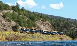A scenic train traveling through a canyon beside a river surrounded by trees and rocky terrain