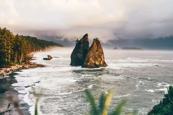 Coastal scene with large rock formations rising out of the ocean, with a sandy beach and forests in the background