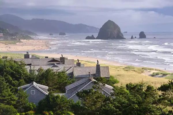 The famous Haystack Rock with an incredible cloudscape building up on the horizon. The mist of the waves spraying up in front while all kinds of birds are flying around the nature reserve. Lots of people down at the Beach enjoying this wonderful summer day.