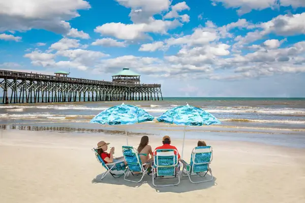 Family relaxing on the beautiful beach, People enjoying summer vacation by the ocean. Family sitting under beach umbrella. Cloudy sky and pier in the background. Folly Beach, South Carolina USA.