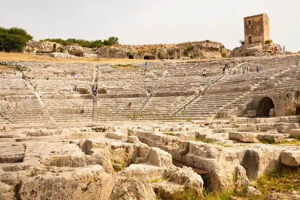 Ancient stone theater ruins with scattered visitors exploring its steps and a tall structure in the background
