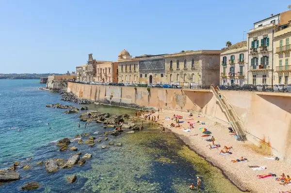 A beach area alongside historic buildings and a coastal wall people enjoying the water and sunbathing