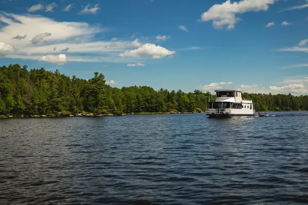 A boat navigating a serene body of water bordered by dense green trees, with a blue sky and scattered clouds overhead