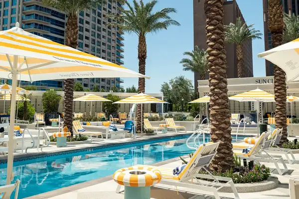 Outdoor lounge area with pool, deck chairs, and umbrellas surrounded by palm trees and buildings