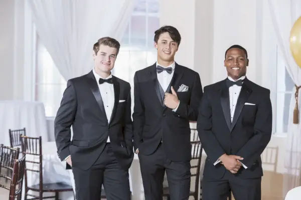 a group of three multi ethnic young men wearing tuxedos, standing in a row, smiling at the camera they are at a party, prom, a wedding or some other special formal event
