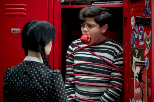 a girl looks at boy tied up in locker with apple in his mouth