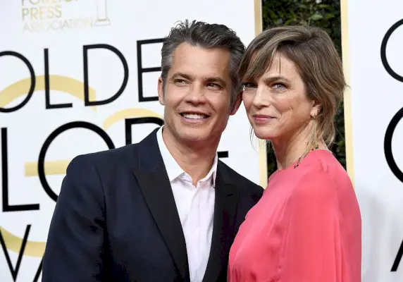 olyphant in navy suit and white button up, knief in red silk dress, pose on the red carpet at nbc