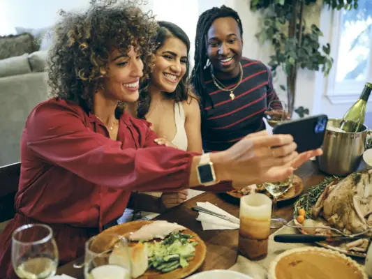 three women taking a selfie over a dinner table with a bucket with wine, turkey, a pumpkin pie, a candle, plates with food on them and a wine glass on the table