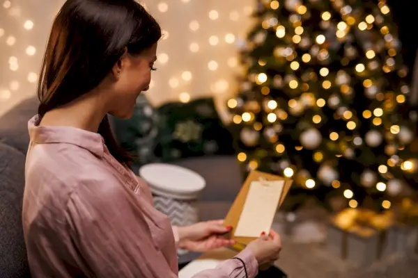 happy young woman sitting on the sofa, by the glistening christmas tree, opening and reading a christmas card she got with her present