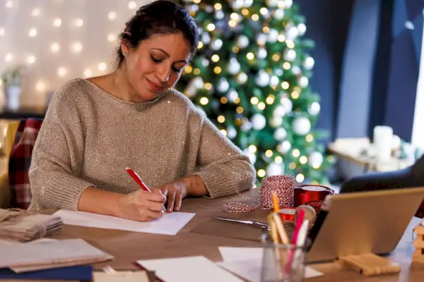portrait of happy mid adult woman sitting at the table, in front of a glistening christmas tree, smiling while writing christmas cards and letters for loved ones