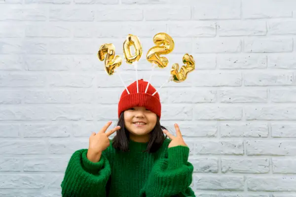 happy asian baby girl in a green sweater close up with golden balloons with numbers 2023 in a hat copy space, white brick wall background, girl looking at the camera, smiling
