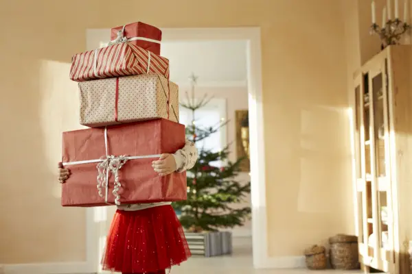 little girl holding tall stack of christmas presents, standing in living room
