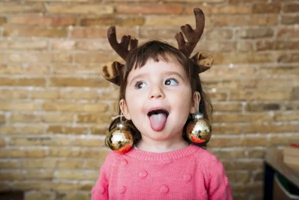 portrait of toddler girl sticking out tongue wearing reindeer antlers headband and christmas baubles