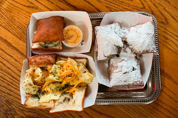 A tray with beignets covered in powdered sugar and sandwiches in white paper containers